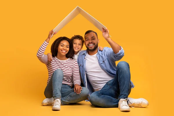 Family Insurance. Happy Black Family Of Three Holding Symbolic Carton Roof Above Heads, Young African American Parents And Preteen Son Posing Together Over Yellow Background In Studio, Copy Space