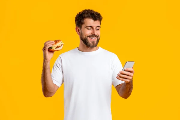 Vrolijke Jongeman Die Hamburger Vasthoudt Met Behulp Van Mobiele Telefoon — Stockfoto