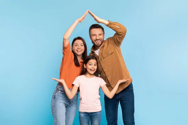 Family insurance. Excited daddy and mommy making symbolic roof of hands above their kid daughter, smiling to camera, celebrating real estate purchase against blue backdrop in studio