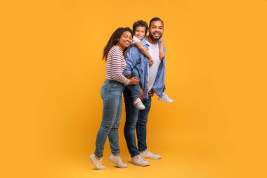 Üç çocuklu mutlu Afrikalı Amerikalı ailenin portresi, kameraya sarılıp gülümseyen, neşeli siyahi ebeveynler ve sarı stüdyonun arka planına karşı eğlenen erkek çocukları.