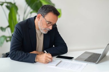 Gözlüklü, üst düzey Kafkasyalı profesyonel ve takım elbiseli biri modern beyaz bir masada dizüstü bilgisayar ve akıllı telefonla çalışırken özenle not alıyor. İş, iş.