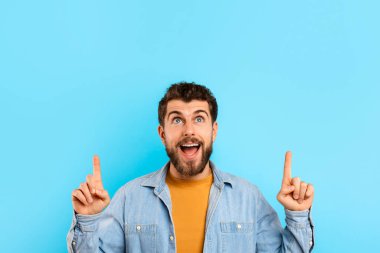Heyecanlı genç adamın portresi, mavi stüdyo arka planına karşı özel indirimli poz vererek yukarı doğru işaret ediyor. Reklam için şablon, tanıtım metni için yeterli kopya alanı sunma