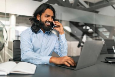 Mutlu yakışıklı doğu iş adamı müşteriyle telefon görüşmesi yapıyor, ofiste çalışıyor, dizüstü bilgisayarda klavye çalıyor, kopyalama alanı kullanıyor. Modern iş konsepti