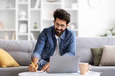 Evde dizüstü bilgisayarın yanına notlar yazan gülümseyen Hintli bir adam mutlu bir doğulu adam çalışıyor ya da internetten çalışıyor. Gri koltukta rahatça oturuyor.