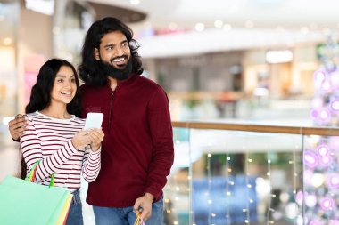 Alışveriş merkezinde alışveriş yaparken internetten promosyon kuponlarını kontrol eden Hintli çift. Neşeli doğulu adam kız arkadaşına sarılıyor, birlikte zaman geçiriyor, mal alıyor, fotokopi çekiyor.