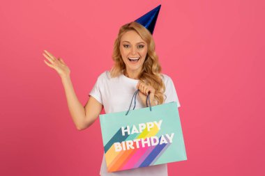 Neşeli bir doğum günü kutlaması. Parti şapkalı gülümseyen sarışın kadın, elinde mutlu yıllar çantası, kameraya bakıyor, sürpriz hediyeler veriyor ve sevinçle kolunu kaldırıyor, pembe stüdyo arka planında.