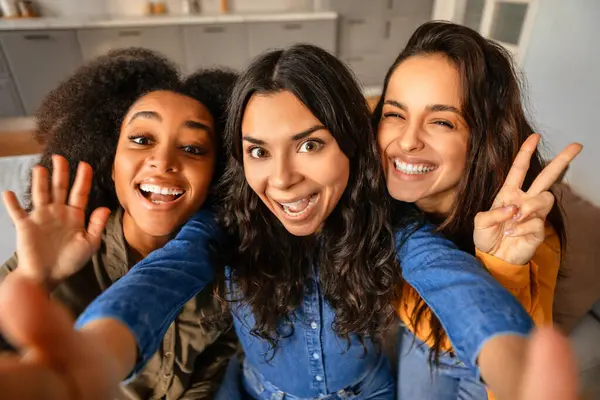 Three joyful young multicultural women taking playful selfie, waving and showing peace signs, as they posing together, making social media content at home. Diverse beauty and friendship
