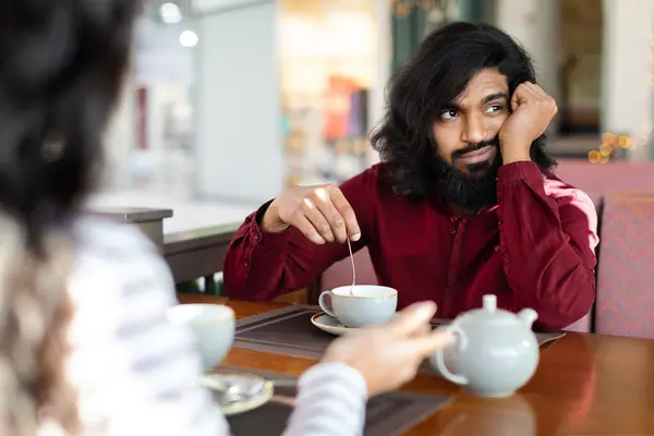 Young bored indian man sitting at table in front of his date or girlfriend and drinking coffee at cozy cafe, looking away. Speed dating, unsuccessful meeting