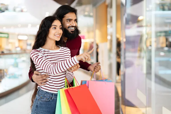 ショッピングモールに立っている若い東部のカップルを愛する買い物バッグ 店の窓を指す 広告のための空白のスペース 消費者主義 季節割引 ショッピングコンセプト — ストック写真