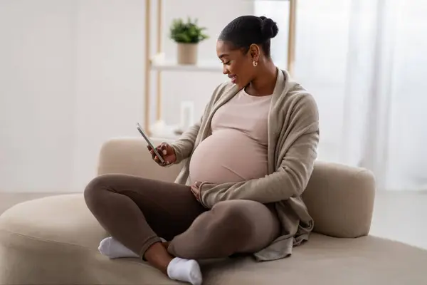 快乐的非洲裔美国年轻孕妇 穿着休闲装 用电话 坐在沙发上 为未来的妈妈们检查漂亮的手机申请 复制空间 — 图库照片