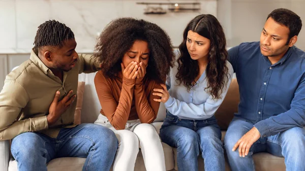 Diverse group of friends providing support to unhappy crying black lady during challenging times, sitting on sofa in modern living room, showcasing strength of true friendship. Panorama