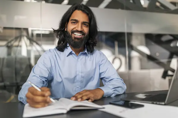 Positive bearded young indian man in formal employee working at modern office, sitting at desk with computer, taking notes in notebook, getting ready for presentation, online meeting, copy space