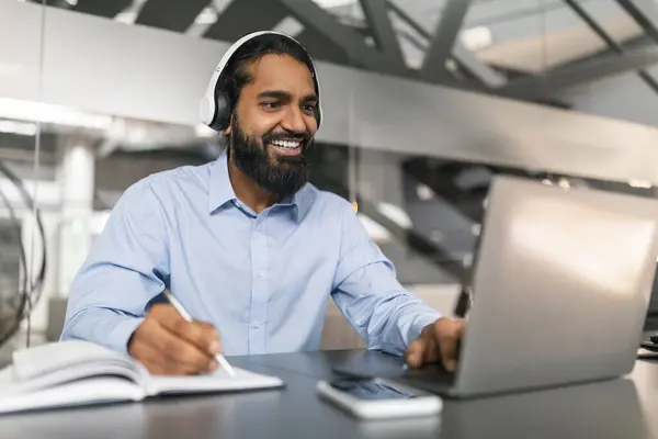 Young indian man having online training or attending webinar, sitting at workdesk at office, using laptop and headset, taking notes and smiling, copy space. Professional online education concept