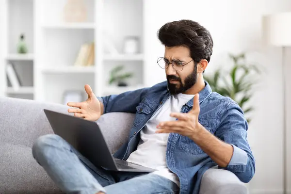 Confused Young Indian Guy Suffering Problems While Using Laptop At Home, Stressed Eastern Man Looking At Computer Screen And Emotionally Gesturing While Sitting On Couch In Living Room, Closeup
