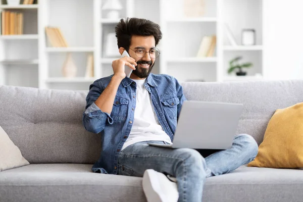 Yakışıklı genç Hintli serbest bilgisayarcı adamın portresi dizüstü bilgisayar kullanıyor ve evde koltukta otururken cep telefonuyla konuşuyor gülümseyen doğulu adam bilgisayarda çalışıyor ve telefonla konuşuyor.