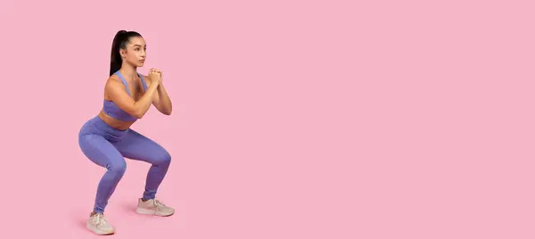 Konzentrierte Junge Frau Kniebeuge Koordiniertem Lila Sportoutfit Vor Pastellrosa Hintergrund — Stockfoto