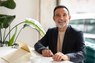 Sempatik, sakallı, kazak üzerine ceket giyen, elinde defter ve fincanla modern bir kafede otururken kameraya gülümseyen beyaz bir adam.