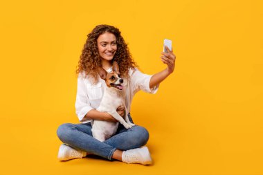 Kıvırcık saçlı kadın, heyecanlı Jack Russell Terrier 'iyle mutlu bir şekilde selfie anı yakalıyor. Parlak sarı arka planda oturuyor.