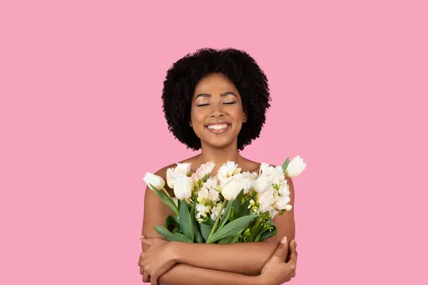 心满意足的年轻非洲裔美国女人闭上双眼 淡淡的微笑 拥抱着一束郁郁葱葱的白色郁金香 在柔和的粉红背景下展现出宁静和幸福 — 图库照片