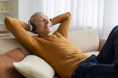 Keyifli gri saçlı adam rahat oturma odasında kanepeye yaslanıyor, kablosuz kulaklık kullanıyor, kapalı gözlerle müzik dinliyor, dedem evde dinleniyor, boşluğu kopyalıyor.