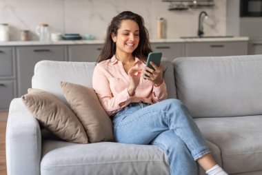 Yumuşak pembe bluzlu ve kot pantolonlu gülümseyen genç bir kadın rahat koltuğunda eğlenirken akıllı telefonuyla iyi ışıklandırılmış odasında mesaj atıyor, internette sörf yapıyor.
