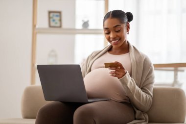 Güzel siyah hamile kadın bebek için internetten alışveriş yapıyor, dizüstü bilgisayar ve kredi kartı kullanıyor, evdeki kanepede oturuyor, boş bir alanda. Gülümseyen Afrikalı bayanın internetten alışveriş yapmasını bekliyor.