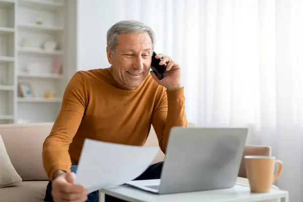 Smiling old man have business call while working from home. Elderly entrepreneur or manager sitting on couch, talking on phone, holding papers, looking at computer screen, copy space