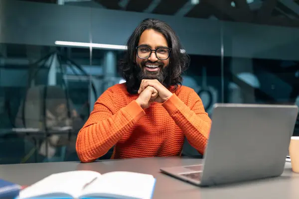 Upbeat professional indian guy with long hair and spectacles, wearing orange pullover, beams while working on his laptop in a contemporary office environment. IT management, tech employees