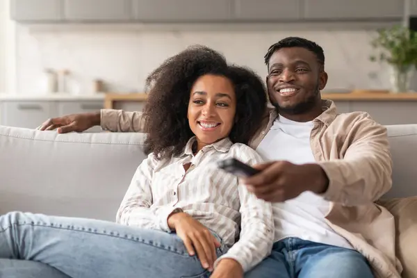 Casual TV time. Smiling African American couple watching television together at home interior, husband holding and pointing remote controller switching television channels. Domestic comfort and fun