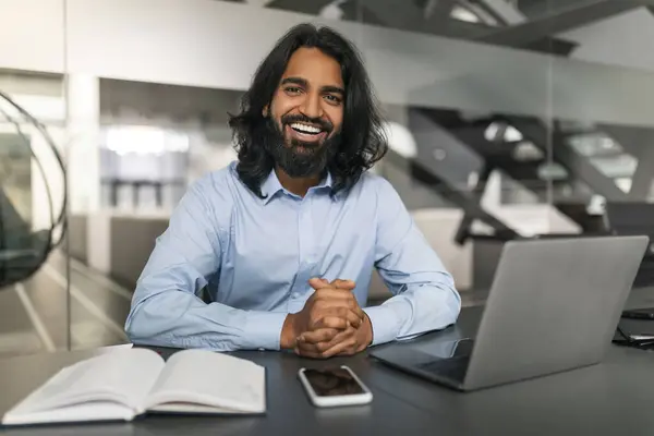 Portrait of professional young indian guy sitting at his desk with laptop pc in modern office. Millennial businessman using computer for online job at workplace, having virtual meeting