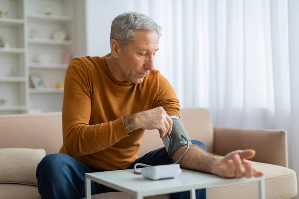 Handsome grey-haired senior man in casual outfit sitting on couch at home, checking blood pressure on his own. Pensioner using modern tonometer, experiencing hypertension, copy space