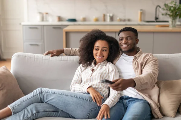Home entertainment. Black millennial couple watching TV, husband and wife enjoy relaxing weekend together, switching channels with remote controller in their cozy living room