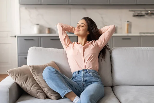 Serene woman in pink blouse and jeans sitting back with her hands behind her head on comfortable couch, embodying relaxation and ease, free space