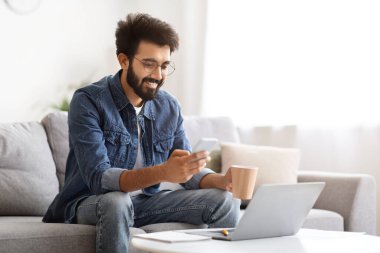 İçerik Hintli adam akıllı telefonunu karıştırıyor ve elinde kahve fincanı tutuyor, doğu kanepede oturan gülümseyen adam modern oturma odasının iç kısmında dizüstü bilgisayarla oturuyor, internet iletişiminin tadını çıkarıyor, uzayı kopyalıyor.