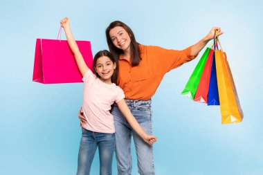 Heyecanlı genç anne ve kız alışverişlerini alışveriş poşetlerinde tutuyorlar, mavi stüdyo arka planında sevinçle poz veriyorlar. Mevsimlik satışlar ve indirimler, Kara Cuma teklifi