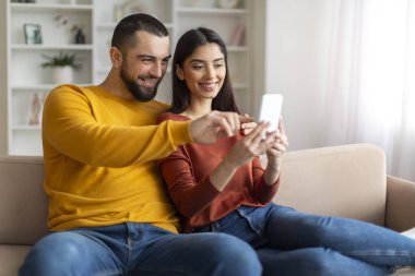 Mutlu genç adam ve kadın birlikte kanepede dinlenirken akıllı telefon kullanıyor, evli beyaz çift sarılıyor ve cep telefonu ekranına bakıyor, en yeni cep telefonu uygulamasını kullanarak gülümsüyor.