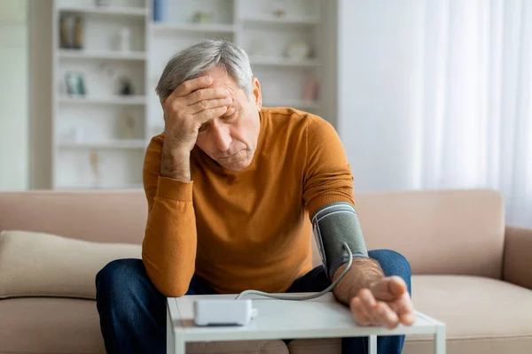 Unhealthy senior man using an upper arm blood pressure monitor while sitting at table in living room, elderly gentleman feeling unwell at home, looking at device, having problems with health