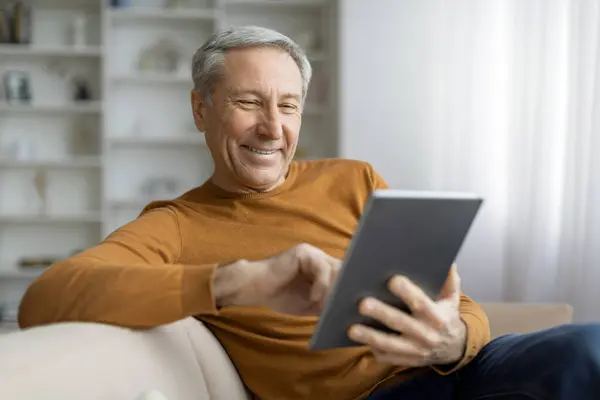 Happy handsome senior man chilling alone at home, sitting on couch in cozy living room interior, using digital tablet, read news online, websurfing, copy space. Digital world in senior age