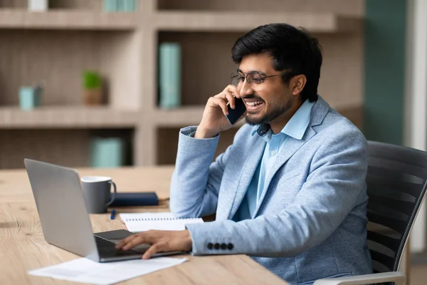 Successful Indian businessman engaging in conversation on his mobile phone while working online on laptop, showcasing effective communication and positive work atmosphere at office