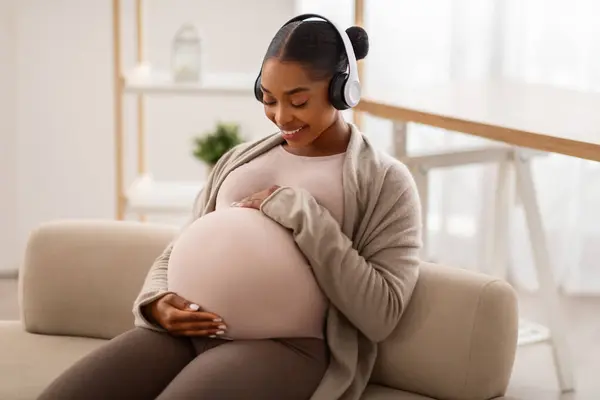 Classic music and influence on baby during pregnancy. Positive expecting young black woman enjoying music at home, sitting on couch, using wireless headphones, embracing her big tummy