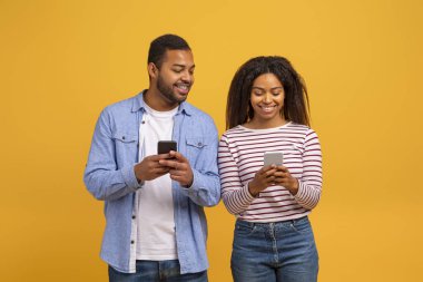 Zeki telefonlarını kullanan neşeli genç siyah çift, mutlu Afrikalı Amerikalı erkek ve kadın sıcak gülümsemeyle dijital bağlantı anlarını paylaşıyor, sarı stüdyo arka planına karşı duruyorlar.