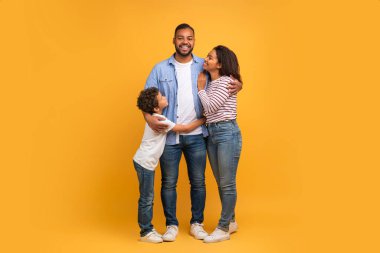 Aile sıcaklığı. Afrikalı Amerikalı baba, anne ve oğul, sarı arka plana sarılıyorlar, mutlu siyah ebeveynler ve gülümseyen ve kameraya bakan çocuk, fotokopi aletiyle uzun bir çekim.