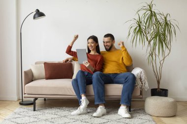 Mutlu genç çift evde dijital tabletle başarıyı kutluyor, mutlu beyaz karı koca internette iyi haberler veriyor, parlak oturma odasındaki bej koltukta oturuyor, boşluğu kopyalıyor.