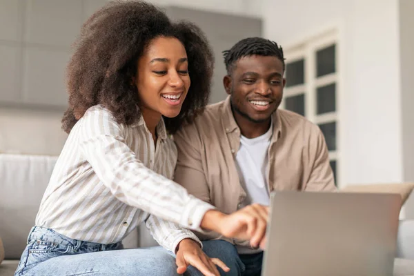 Weekend tech bonding. Young black couple smiling while using laptop on sofa together, woman pointing at screen, shopping and showing webstore website to husband in modern living room at home