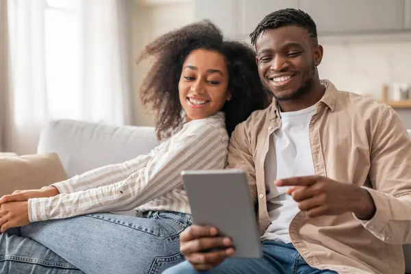 Happy African American couple with digital tablet enjoys online content together, man pointing at computer showing funny video to wife, sitting on sofa in modern living room at home