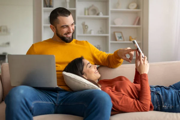 Happy Young Couple Reading Message On Smartphone And Using Laptop At Home, Cheerful Millennial Spouses Browsing New App On Mobile Phone While Relaxing On Couch, Woman Lying On Husbands Lap