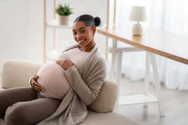 Maternity, pregnancy concept. Beautiful young african american pregnant woman wearing casual clothing sitting on couch at home, embracing her big belly, smiling at camera, copy space