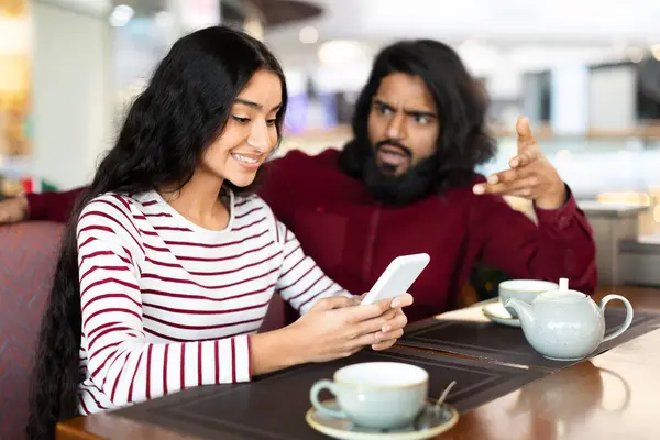 Angry young indian man gesturing and yelling, have fight with girlfriend smiling at smartphone screen while romantic date at coffee shop. Eastern couple experiencing difficulties in relationships