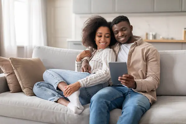 Connected couple. Happy black husband and wife enjoying movie on tablet sitting on sofa, relaxing on weekend in living room interior. Modern lifestyle, digital entertainment at home