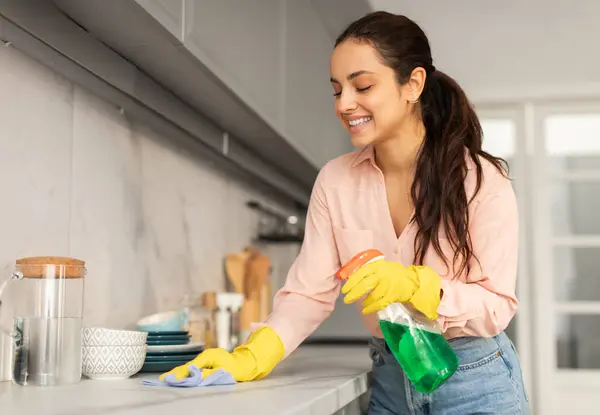 Smiling woman in blouse and yellow gloves, happily wiping the kitchen countertop with blue cloth and green spray bottle in hand, free space